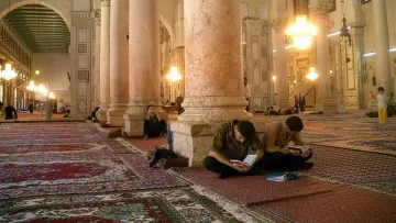 L'image montre l'intérieur d'une grande mosquée, caractérisée par de hauts plafonds et de majestueuses colonnes. Le sol est recouvert de tapis, et plusieurs personnes sont assises sur le sol, certaines concentrées sur leurs ordinateurs portables, tandis que d'autres semblent prier ou méditer. La lumière naturelle entre par les fenêtres, créant une ambiance calme et sereine. Des lustres pendent du plafond, ajoutant une touche d'élégance à cet espace sacré.