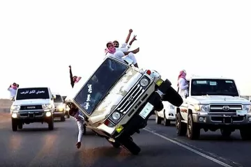 L'image montre plusieurs véhicules tout-terrain sur une route. Un des véhicules est en train de faire un équilibrage sur deux roues, avec des personnes à l'intérieur et à l'extérieur qui semblent célébrer et s'amuser. Les passagers portent des vêtements traditionnels, et l'ambiance générale semble festive. En arrière-plan, d'autres véhicules sont visibles, créant une scène dynamique et animée.