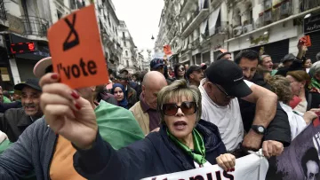 The image depicts a crowd of protesters gathered in an urban setting, likely during a demonstration. A woman in the foreground is holding an orange sign that reads "l'vote" while wearing sunglasses and a green top. The crowd behind her appears to be a mix of men and women, some holding signs and banners. The atmosphere conveys a sense of urgency and solidarity among the protesters, who seem to be advocating for a political cause. The background includes urban architecture, suggesting a city environment.