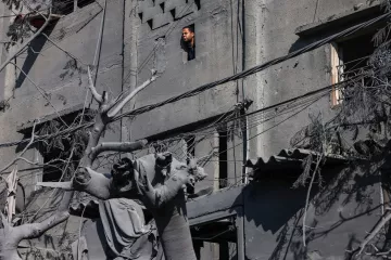 L'immagine mostra un edificio gravemente danneggiato, con i muri di cemento visibilmente rovinati. Da una finestra rotta, si intravede una persona che osserva l'esterno, mentre nel primo piano si trova una statua, anch'essa coperta di polvere e detriti. Le linee elettriche pendono disordinatamente, e l'atmosfera è cupa a causa dei colori grigi e della desolazione del contesto. La scena sembra suggerire un ambiente colpito da un evento catastrofico, con evidenti segni di distruzione e abbandono.