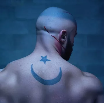 The image depicts a muscular, bald man from the back, showcasing a prominent tattoo on his back. The tattoo features a crescent moon and a star, which are often associated with various cultural and religious symbols. There are visible scars or cuts on his scalp and a streak of blood running down, suggesting a recent injury or intense situation. The background has a dark, moody atmosphere, possibly indicating a gritty or dramatic context.