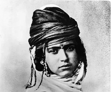 L'image montre un portrait noir et blanc d'une personne portant un turban orné et des bijoux. Le regard de la personne est intense, et son expression transmet une certaine dignité. Elle est vêtue de vêtements traditionnels qui reflètent une culture spécifique. Les détails du turban et des ornements ajoutent une dimension riche et texturée à l'image.
