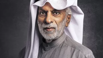 L'image montre un homme âgé avec une barbe grise, portant un vêtement traditionnel de style arabe, typiquement un thobe gris avec des rayures. Il a également un agal noir et un ghutrah blanche, qui est un couvre-chef traditionnel. L'arrière-plan est sombre, ce qui met en valeur son expression sage et son apparence traditionnelle.