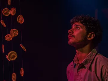 L'image montre un jeune homme regardant avec curiosité quelque chose au-dessus de lui. Il est entouré d'une ambiance sombre, avec des éclairages qui créent des reflets colorés, probablement des nuances de bleu et de rose. Des rondelles d'orange sèchées pendent de fil, créant un effet visuel intéressant, tandis que l'expression du jeune homme semble pensive et captivée par la scène.