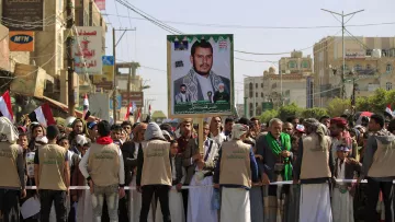 تظهر في الصورة مجموعة من الأشخاص مجتمعين في مظاهرة أو تجمّع شعبي. في الخلفية، يُرفع لافتة كبيرة تحمل صورة لأحد الشخصيات، ويبدو أن المتظاهرين يرتدون ملابس متشابهة. تعكس الأعلام الموجودة في الصورة انتماءات وطنية، كما يظهر أن هناك تفاعلًا وحماسًا بين المشاركين. الجو يعكس نوعًا من الاحتشاد والتضامن حول قضية معينة.