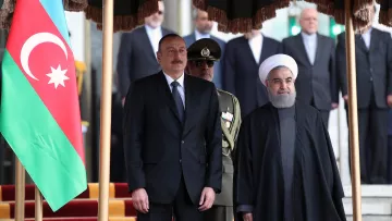 En la imagen se pueden ver a dos líderes de países que están de pie juntos. A la izquierda, hay una bandera de Azerbaiyán. Uno de los líderes lleva un traje negro y corbata, mientras que el otro viste una indumentaria tradicional oscura. Detrás de ellos, hay un grupo de personas que parecen ser miembros de la seguridad o funcionarios, y en el fondo se observan algunos edificios. La escena parece ser un acto formal o una reunión oficial.
