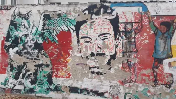 L'image montre une fresque murale riche en couleurs et en détails. À gauche, on peut voir un personnage avec des ailes et un visage expressif, qui semble être une représentation stylisée d'une créature. À droite, se trouve un visage humain qui attire l'attention, entouré de motifs vibrants. En arrière-plan, divers éléments graphiques et des inscriptions en arabe ajoutent une dimension culturelle. L'ensemble donne une impression de dynamisme et de créativité, typique de l'art urbain.