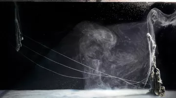 The image showcases a mesmerizing underwater scene featuring swirling currents and bubbles. The foreground has a smooth, almost glass-like surface with a hint of foam, while the background features abstract formations that resemble smoke or flowing water. Thin, delicate tendrils or strings extend across the scene, creating an ethereal atmosphere. The overall color palette is dominated by dark tones, emphasizing the luminous qualities of the water and creating a sense of depth and motion.
