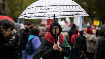 En la imagen, se puede ver a una persona que sostiene un paraguas con la palabra "Palestine" escrita en él. La persona lleva una bandera de Palestina sobre los hombros y parece estar en una manifestación o protesta. Alrededor se pueden observar otras personas, algunas con paraguas y banderas, en un ambiente cargado de emociones y solidaridad. El clima parece nublado o lluvioso, lo que añade a la atmósfera del evento.