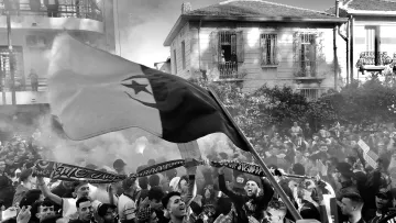 The image depicts a large crowd gathered in an urban setting, likely during a festive or protest event. People are seen waving flags and holding scarves, creating a vibrant atmosphere. Smoke is rising in the air, possibly from flares or fireworks, adding to the intensity of the scene. The buildings in the background have a mix of architectural styles, hinting at a lively neighborhood. The overall mood appears energetic and spirited, with individuals showing strong emotions and camaraderie.