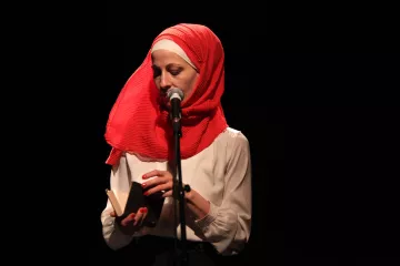 L'image montre une femme debout devant un microphone sur scène. Elle porte un hijab rouge vif et une blouse blanche. Elle tient un petit livre dans ses mains, semblant s'apprêter à lire ou à partager un texte. L'éclairage est sombre, mettant en valeur son visage et l'objet qu'elle tient. L'atmosphère semble intime et concentrée.