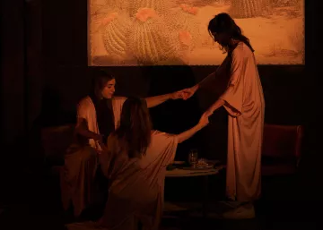 L'image présente une scène théâtrale ou artistique, où trois femmes en vêtements fluides de couleur beige se tiennent autour d'une table basse. Elles semblent engagées dans une interaction attentive, tenant les mains les unes des autres. La lumière est tamisée, créant une atmosphère intime et mystérieuse. En arrière-plan, il y a un tableau représentant des cactus dans un paysage désertique, ajoutant une touche de nature à la scène. Les expressions des femmes ne sont pas clairement visibles, contribuant à l'ambiance intrigante de l'image.
