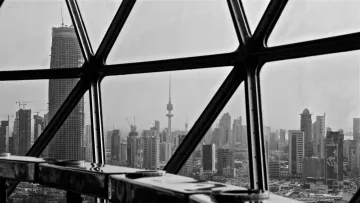 L'image montre une vue urbaine à travers une grande fenêtre en verre avec un cadre géométrique. Au premier plan, on aperçoit une surface réfléchissante et des éléments architecturaux. À l'extérieur, on découvre un paysage de ville en noir et blanc, avec des gratte-ciels modernes et une tour emblématique qui se dresse au loin, probablement un symbole de la skyline. La scène dégage une atmosphère à la fois contemporaine et dynamique.