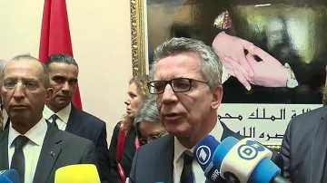 L'image montre un groupe de personnes lors d'une conférence de presse ou d'un événement officiel. Au premier plan, un homme s'exprime devant des caméras et des micros, entouré de plusieurs autres individus qui semblent attentifs. En arrière-plan, on peut voir un portrait ou une image représentant des mains avec des symboles significatifs, ainsi qu'un drapeau. L'atmosphère semble formelle, probablement liée à des discussions d'ordre politique ou diplomatique.