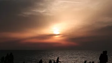 L'immagine mostra una scena al tramonto sulla spiaggia. Il cielo è dipinto di tonalità calde, con sfumature di arancione e rosa, mentre il sole si avvicina all'orizzonte. In primo piano, si vedono diverse silhouette di persone che camminano lungo la riva, alcune sedute o accoccolate sulla sabbia. La superficie dell'acqua riflette i colori del cielo, creando un'atmosfera calma e suggestiva.