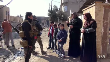 L'image montre une scène dans une rue dévastée, probablement dans une zone de conflit. Un soldat armé est debout, en uniforme militaire, tenant son arme. Il semble interagir avec deux femmes voilées qui lui parlent. À proximité, plusieurs enfants, dont deux garçons, regardent la scène. Le décor est marqué par des bâtiments endommagés et des débris qui témoignent d'une situation difficile. L'atmosphère semble tendue mais il y a aussi une interaction sociale entre les personnes présentes.