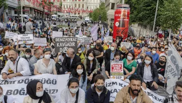 L'image montre une manifestation en plein air avec de nombreuses personnes rassemblées dans une rue. Les manifestants portent des masques et brandissent des pancartes avec des slogans. L'atmosphère semble mobilisée, et il y a beaucoup de drapeaux en arrière-plan. Les participants sont assis pour exprimer leur message, signalant une volonté de protester pacifiquement.