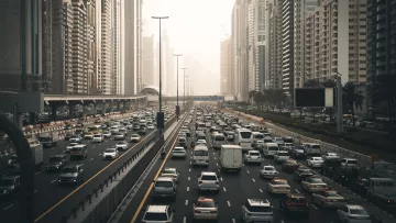 L'image montre une vue d'une route très fréquentée dans une grande ville moderne, entourée de gratte-ciel. On peut voir de nombreux véhicules qui circulent dans les deux sens, créant un embouteillage. La lumière du soleil est tamisée, donnant une atmosphère légèrement brumeuse, peut-être due à la pollution ou à la météo. Les bâtiments en arrière-plan sont imposants et reflètent l'architecture contemporaine, avec des façades en verre et en béton. Des palmiers sont visibles sur le côté de la route, ajoutant une touche de verdure à ce paysage urbain.
