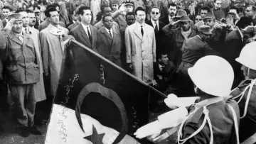 The image depicts a historical scene with a large crowd gathered, likely for a ceremonial or significant event. In the foreground, two soldiers are saluting while holding a flag, which features a crescent and a star, elements often associated with some national flags. The crowd appears to consist of men in various types of clothing, with some wearing military uniforms and others in suits. The overall atmosphere suggests a moment of national importance, possibly related to independence or a political event.