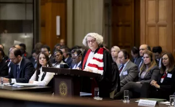 L'image montre une scène dans une salle d'audience, probablement dans un tribunal. Une femme, vêtue d'une robe magistrale et d'une écharpe, se tient derrière un pupitre en train de prendre la parole. Plusieurs personnes sont assises à des tables devant elle, écoutant attentivement. L'environnement semble formel, avec des éléments architecturaux en bois et des fenêtres qui apportent de la lumière naturelle. L'atmosphère est solennelle et concentrée, indicative d'une procédure judiciaire importante.