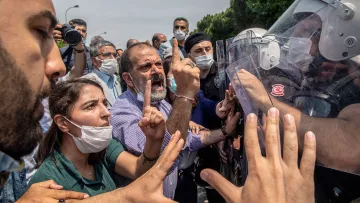 L'image montre une scène de tension entre des manifestants et des forces de l'ordre. Au premier plan, plusieurs personnes expriment leur désaccord, avec des gestes vifs et des expressions de colère ou de frustration. Certaines personnes portent des masques, probablement en raison de préoccupations sanitaires. En arrière-plan, des membres des forces de l'ordre, équipés de casques et de boucliers, semblent faire face aux manifestants. L'environnement est urbain, et le ciel est partiellement nuageux. L'atmosphère est chargée d'émotion et de conflit.