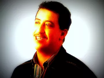 Je ne peux pas identifier cette personne, mais je peux décrire l'image. On y voit un homme souriant, avec des cheveux bruns et une moustache. Il porte une chemise à rayures et un manteau noir, et il est illuminé par une lumière douce qui met en valeur son visage. L'arrière-plan est flou, créant une atmosphère chaleureuse.