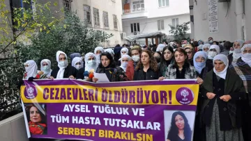 En la imagen, un grupo de personas se encuentra reunido en una manifestación. Hay mujeres con pañuelos en la cabeza, y algunas de ellas sostienen pancartas que expresan mensajes en apoyo a Aysel Tuğluk y a otros prisioneros enfermos. El ambiente parece ser de protesta, con un enfoque en la liberación de detenidos. Se observa una edificación al fondo, que sugiere que la manifestación se lleva a cabo frente a un edificio público o institucional.