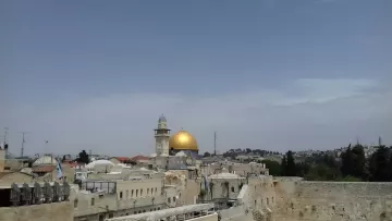 L'immagine mostra una vista panoramica di una città con una predominante cupola dorata, che è probabilmente il Dôme della Roccia, un noto sito religioso a Gerusalemme. Intorno ci sono edifici con tetti piatti e una torre, mentre il cielo appare sereno e leggermente nuvoloso. Gli alberi e le colline sullo sfondo aggiungono profondità alla scena.