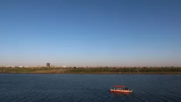 The image depicts a serene river landscape under a clear blue sky. In the foreground, a small boat with a red canopy is floating on the water, which reflects the sky. In the background, there is a sandy shore lined with greenery, and a distant cityscape can be seen, with buildings rising against the horizon. The overall scene conveys a peaceful and picturesque atmosphere.