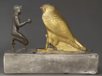 L'image représente une sculpture en métal, où l'on peut voir un personnage humain à genoux tenant un objet dans ses mains, face à un grand hibou doré. Le personnage est sculpté de manière détaillée, avec des traits qui évoquent un style ancien, probablement lié à l'Égypte ou à une culture similaire. Le hibou, de couleur dorée, symbolise peut-être la sagesse ou un aspect divin. La base de la sculpture est en métal argenté, offrant un contraste intéressant avec la brillance du hibou doré. Cette œuvre semble revêtir une signification symbolique ou religieuse.