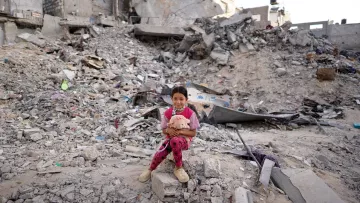 L'immagine ritrae una bambina seduta su un cumulo di macerie, circondata da un paesaggio distrutto. Il suo sguardo è diretto verso la fotocamera, mentre tiene un peluche tra le braccia. Indossa un pigiama a pois rosa e sembra essere in un ambiente devastato, con edifici in rovina sullo sfondo. La scena trasmette una forte emozione, evidenziando la vulnerabilità e la resilienza della giovane ragazza in un contesto difficile.