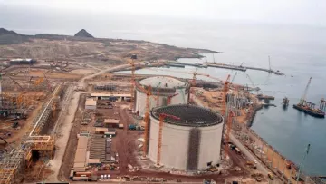 L'image montre un site industriel en construction, probablement une installation de gaz naturel ou une centrale électrique. On peut voir plusieurs grands réservoirs cylindriques, entourés de grues et de matériaux de construction. L'environnement est côtier, avec une vue sur la mer et des montagnes en arrière-plan. Il y a également des routes et des véhicules de construction présents sur le site. Le paysage est principalement désertique et montagneux.