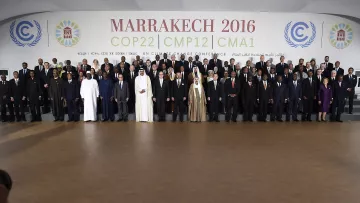L'image montre un groupe de participants rassemblés lors de la conférence COP22 qui s'est tenue à Marrakech en 2016. Les membres représentés incluent des dirigeants et des délégués de divers pays, vêtus de costumes formels et de tenues traditionnelles. Ils se tiennent debout sur des gradins, souriant pour la photo officielle, avec un fond affichant le nom de l'événement et des logos des Nations Unies. L'ambiance évoque un sentiment de collaboration internationale pour faire face aux défis climatiques.
