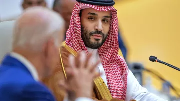 L'image montre un homme portant une tenue traditionnelle saoudienne, comprenant un thobe et un ghutrah (un couvre-chef en tissu). Il est assis à une table lors d'une réunion, fixant le spectateur avec un regard sérieux. À l'arrière-plan, un autre homme, dont le visage est partiellement visible, semble être en train de parler ou de poser une question. L'environnement suggère un cadre formel et officiel, comme une conférence ou un sommet.