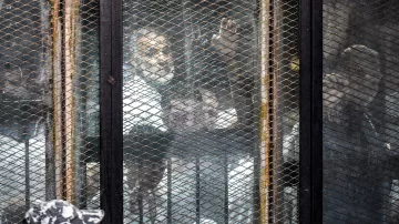 L'image montre une scène où des personnes se trouvent derrière une grille ou une clôture métallique. On aperçoit plusieurs individus, probablement en détention, qui semblent observés par des gardiens ou des forces de l'ordre en uniforme. L'atmosphère semble tendue, et il y a un mélange de visages. L'environnement donne une impression de confinement et de surveillance.