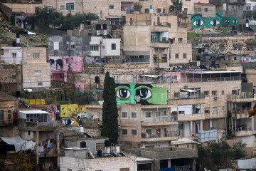 L'image montre un paysage urbain d'une ville, où plusieurs bâtiments sont alignés sur une colline. À travers ces constructions, on peut voir de grandes peintures murales représentant des yeux, aux couleurs vives, qui ajoutent une dimension artistique à cet environnement. Les bâtiments varient en couleurs et en textures, allant du beige au rose, et certains semblent en cours de rénovation. L'atmosphère générale évoque un mélange de quotidien et de créativité, avec une forte présence d'art urbain.