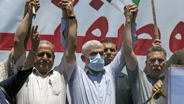 الصورة تُظهر مجموعة من الرجال في تجمع حاشد، حيث يرفع بعضهم أيديهم في إشارة للتضامن أو الاحتجاج. يرتدي أحد الأشخاص في المنتصف قناعاً للوجه، مما يشير إلى أنه قد يكون في سياق صحي معين. خلفهم يوجد لافتة كبيرة مكتوب عليها نص باللون الأحمر. الجو العام يوحي بالتعبير عن موقف أو رسالة معينة.