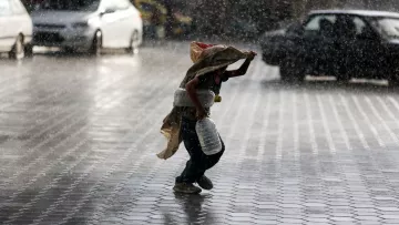 Nell'immagine si vede un bambino che corre sotto la pioggia intensa. Il ragazzo tiene un grande contenitore in una mano e si protegge dalla pioggia con un pezzo di stoffa. Sullo sfondo, ci sono diverse auto e una strada bagnata, mentre le gocce d'acqua creano un'atmosfera drammatica e vivace. La scena trasmette una sensazione di urgenza e resilienza, evidenziando il contrasto tra il bambino e le avversità del tempo.