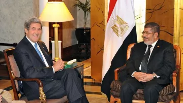 L'image montre deux hommes assis dans un cadre formel. L'un d'eux porte un costume sombre et sourit, tandis que l'autre, également en costume, a un sourire amical. En arrière-plan, on peut voir un drapeau égyptien ainsi qu'un éclairage chaleureux et des éléments de décoration élégants. L'atmosphère semble détendue et cordiale, suggérant une rencontre diplomatique.