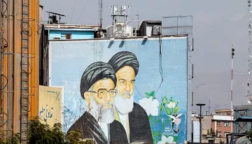 L'image montre une grande fresque murale représentant deux hommes avec des turbans noirs et des barbes blanches. Ils sont peints sur un mur d'un bâtiment, entourés de fleurs et de motifs décoratifs. En arrière-plan, on aperçoit des structures urbaines et un ciel légèrement nuageux. Cette fresque semble symboliser une figure ou un message important dans le contexte culturel ou historique de la région.