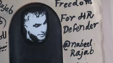 L'image représente un graffiti sur un mur. On y voit un portrait en noir et blanc d'un homme, accompagné d'un texte écrit en anglais. Le message indique "Freedom For HR Defender" (Liberté pour le défenseur des droits de l'homme) et mentionne un nom d'utilisateur Twitter "@nabeelrajab". Le style artistique est simple, avec un fond sombre et des lettres noires qui ressortent sur la surface claire du mur. Cela semble être une œuvre engagée, soulignant le soutien à la défense des droits humains.