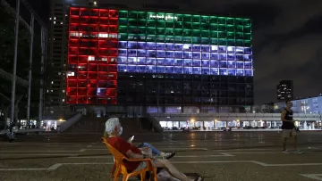The image shows a large building illuminated with colorful lights at night, displaying the colors of a flag: red, green, and white. In the foreground, an elderly man is sitting in a plastic chair, looking at his phone. The scene conveys a sense of tranquility, with the illuminated building serving as a striking backdrop in a public square. In the background, other people can be seen walking or engaging in activities. The atmosphere appears calm and reflective.