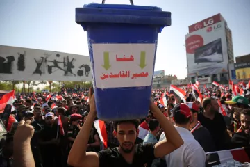 L'image montre une grande manifestation avec une foule rassemblée. Au premier plan, un homme porte un grand seau bleu sur sa tête, qui affiche des inscriptions en arabe. Derrière lui, on peut voir de nombreux drapeaux, probablement ceux de l'Égypte, et des affiches graphiques sur un mur. La scène dégage une atmosphère de protestation, avec des gens mobilisés pour une cause.