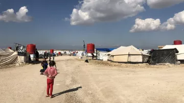 L'image montre un camp de réfugiés avec des tentes disposées de part et d'autre d'un chemin de terre. Au fond, on aperçoit des nuages blancs dans un ciel bleu. Des enfants sont présents, certains semblent jouer ou se déplacer dans le camp. Les abris sont principalement en toile, avec quelques réservoirs d'eau visibles. L'atmosphère paraît calme malgré les conditions de vie apparentes de ces lieux.