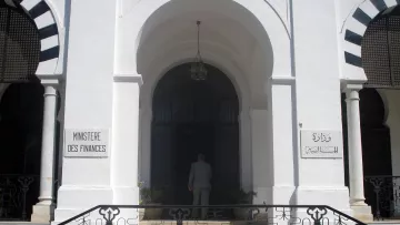 تظهر الصورة مدخل مبنى حكومي يُعرف باسم "وزارة المالية". يتميز المبنى بتصميم معماري جذاب، حيث يمكن رؤية الأبواب الكبيرة والنوافذ المزخرفة. المدخل محاط بأعمدة وأقواس، مما يعطيه طابعاً تقليدياً. هناك لافتات على الجانبين، واحدة باللغة الفرنسية والأخرى بالعربية، تشير إلى اسم الوزارة. يُلاحظ أيضاً شخص يقف أمام المدخل، مما يعكس النشاط في هذا المكان.