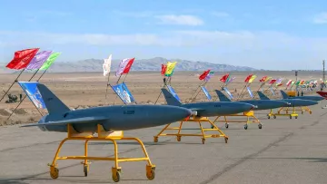 The image depicts a row of military drones lined up on a tarmac. Each drone is mounted on a yellow stand and features colorful flags attached to poles, which are positioned behind them. The background shows a vast, open landscape with distant mountains under a clear sky, suggesting a desert environment. The overall scene conveys a sense of military readiness or demonstration.