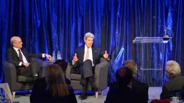 L'image montre une scène de conférence ou de discussion, où deux intervenants sont présents sur une scène. À gauche, un homme est assis sur un fauteuil, écoutant attentivement, tandis qu'à droite, un autre homme parle avec des gestes, probablement en train de partager ses idées ou répondre à des questions. La scène est éclairée avec un fond de rideaux bleus, créant une ambiance professionnelle et formelle. Il y a également un podium avec un microphone, prêt à être utilisé. En bas, on peut apercevoir un public assis, attentif à la conversation.