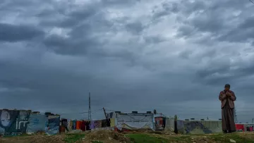 در این تصویر، یک مرد در منطقه‌ای با ساختمان‌های موقتی یا کمپ زندگی می‌کند. آسمان ابری و تیره به نظر می‌رسد و نشان‌دهنده شرایط جوی نامساعد است. در پس‌زمینه، چند چادر یا خانه موقت دیده می‌شود که با پارچه‌ها و وسایل مختلف پوشانده شده‌اند. فضای کلی تصویر حس کمبود و در عین حال استقامت را منتقل می‌کند. مرد به نظر می‌رسد در حال مکالمه یا تفکر است و تصویر کلی فضایی جدی و غم‌انگیز دارد.