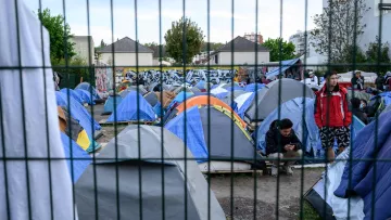 L'image montre un campement de tentes installé dans une zone urbaine. Les tentes sont de différentes tailles et couleurs, principalement en bleu et gris. On peut voir des personnes présentes, certaines debout et d'autres assises, semblant utiliser leur téléphone. En arrière-plan, il y a des bâtiments et un mur décoré de graffitis. L'ensemble du camp est entouré par une clôture, ce qui donne un aspect de confinement à la scène. Les arbres et la végétation ajoutent une touche de verdure au décor.