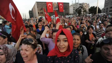 تظهر في الصورة حشود من الناس تجمعوا في مكان واحد، حيث يحمل بعضهم الأعلام التركية. يبدو أن هناك مشاعر حماسية واضحة بين المتظاهرين، حيث يرفعون أصواتهم ويعبرون عن آرائهم. ترتدي بعض النساء الحجاب، بينما تظهر في المقدمة فتاة صغيرة ترتدي غطاء رأس. الخلفية تضم مباني وبعض الصور التي تعكس أجواء الاحتجاج أو الاحتفاء.